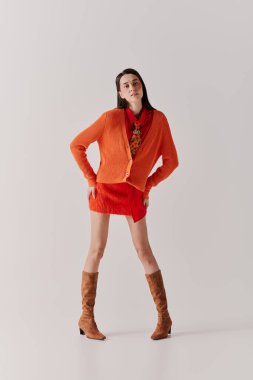 Genç bir kadın kalın turuncu bir hırka ve şık kırmızı bir elbiseyle şık bir şekilde duruyor..