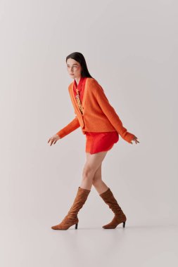 Genç bir kadın rahat turuncu bir süveter ve şık çizmelerle şık bir kıyafetle rahatça yürüyor..