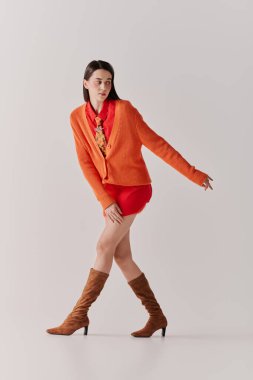 A young woman in an orange cardigan and red outfit strikes a confident pose.