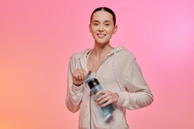 Açık renk kapüşonlu genç bir kadın renkli bir atmosferde su şişesini tutarken gülümsüyor..