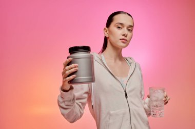 Genç bir kadın, elinde protein kabı ve bir bardak su tutarken şık görünümünü sergiliyor..