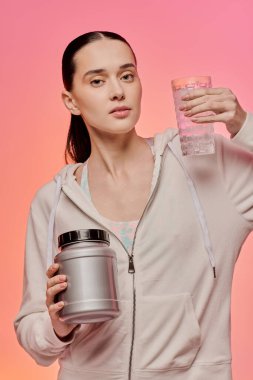 Şık bir kadın, kendine güvenen bir şekilde protein kabıyla poz verir ve parlak bir zemin üzerinde su bulundurur..