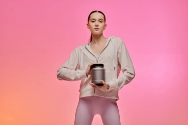 Şık kıyafetli genç bir kadın, yumuşak ışıkta rahatça poz verirken bir fitness konteynırı tutuyor..