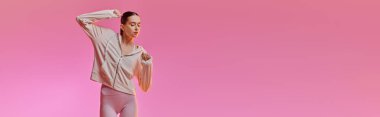 Genç bir kadın parlak pembe bir arka plana karşı şık bir spor elbisesi içinde neşeyle dans ediyor..