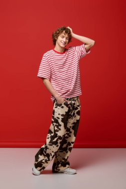 Genç, kıvırcık bir adam modern modayı sergilerken dikkat çekici bir kırmızı stüdyoda güven içinde poz veriyor..