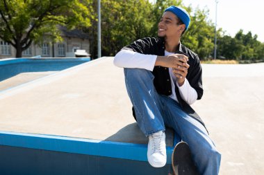 Genç bir adam kaykay parkında rahatça oturur, içkisini yudumlar ve güneşli günün tadını çıkarır..