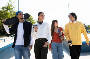 Arkadaşlar kaykay parkında birlikte yürür, gülüşürler ve güneşin altında güzel vakit geçirirler..