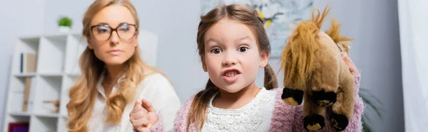 Сердита маленька дівчинка пацієнтка з іграшкою і згорбленим кулаком, що відвідує психолога, банер — стокове фото