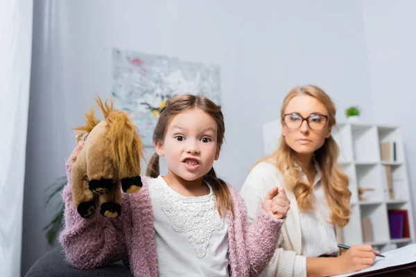 Сердита маленька дівчинка з іграшкою і згорбленим кулаком, що відвідує психолога — стокове фото