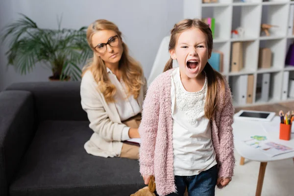 Niña enojada con juguete gritando mientras visita al psicólogo - foto de stock