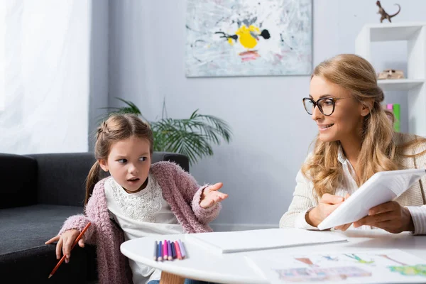 Niña confundida con lápiz que se muestra en la tableta digital mientras visita al psicólogo - foto de stock