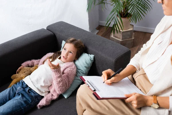 Bambina sdraiata sul divano con giocattolo mentre racconta problema allo psicologo — Foto stock