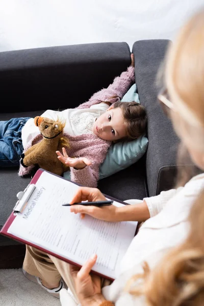 Partial view of psychologist writing in medical history while little girl telling problem — Stock Photo