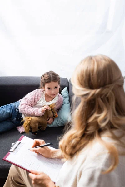 Menina deitada no sofá com brinquedo ao dizer problema para psicólogo — Fotografia de Stock