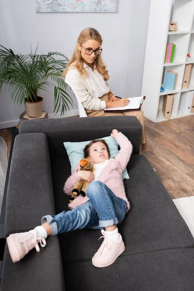 Kleines Mädchen liegt mit Spielzeug auf Sofa und erzählt Problem dem Psychologen — Stockfoto