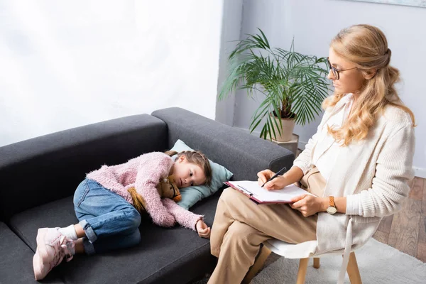 Bambina sdraiata sul divano con giocattolo mentre racconta problema allo psicologo — Foto stock