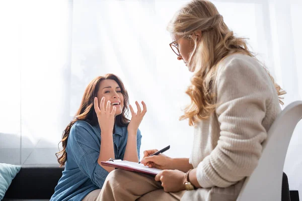 Triste bruna donna gesticolando mentre il terapeuta prende appunti nella storia medica del paziente — Foto stock
