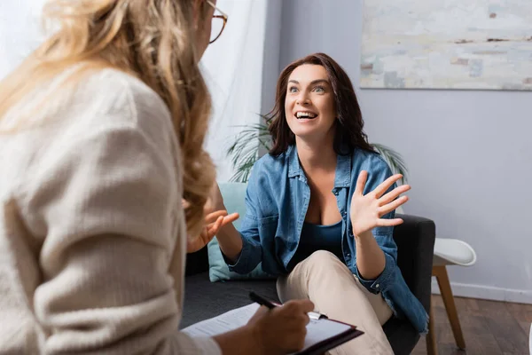 Sorrindo morena mulher gesticulando enquanto terapeuta escrevendo na história médica — Fotografia de Stock