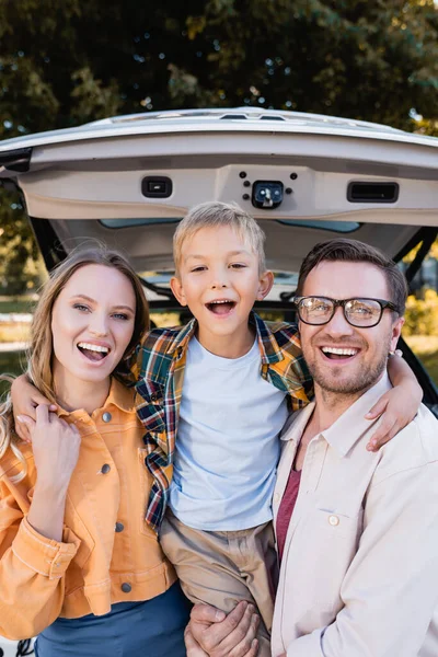 Веселий хлопчик обіймає батьків біля машини під час подорожі на відкритому повітрі — стокове фото