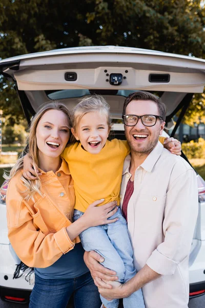 Chica alegre abrazando a los padres cerca del coche durante el viaje al aire libre - foto de stock