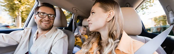 Smiling parents looking at each other near kid on blurred background in car, banner — Stock Photo