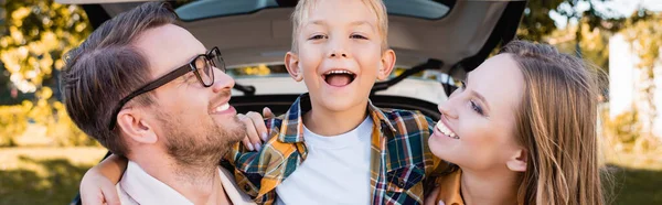 Веселый сын, обнимающий родителей возле машины на размытом фоне на открытом воздухе, баннер — стоковое фото