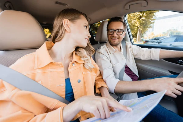 Lächelnder Mann blickt Frau an und zeigt auf Landkarte auf verschwommenem Vordergrund im Auto — Stockfoto