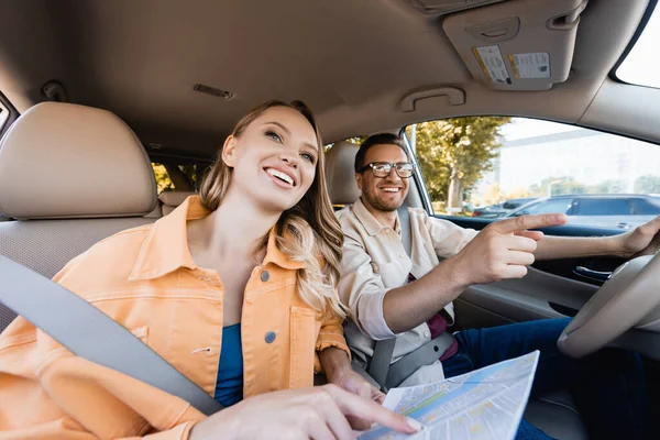 Sonriente hombre apuntando lejos cerca de la esposa con mapa en el coche durante el viaje - foto de stock