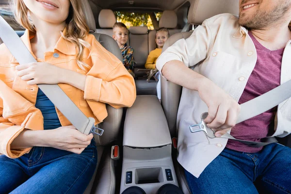 Усміхнені батьки тримають ремені безпеки біля дітей на розмитому тлі в машині — стокове фото