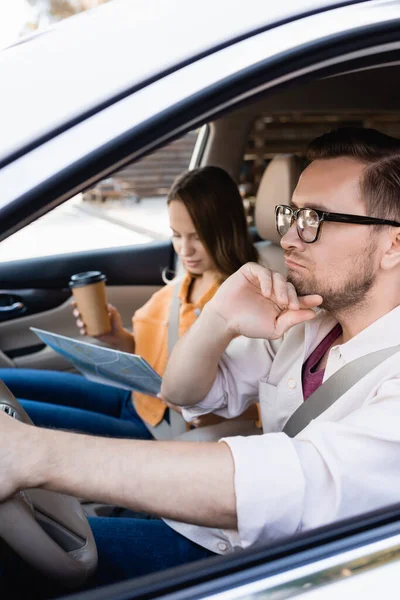 Hombre pensativo conduciendo coche cerca de la esposa con café para ir y mapa sobre fondo borroso - foto de stock