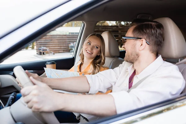 Mujer sonriente con mapa y café para ir mirando al marido conduciendo coche - foto de stock