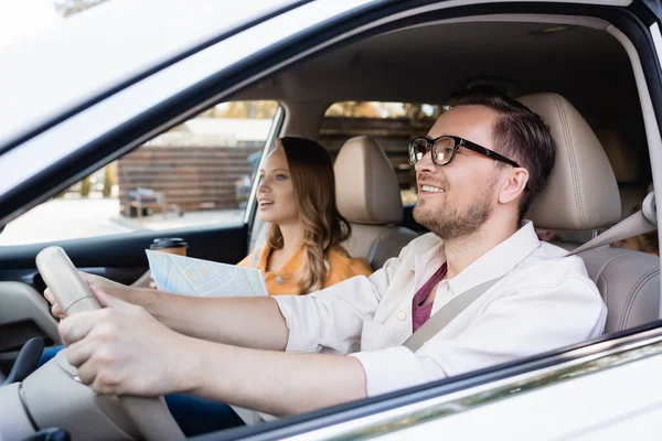 Uomo sorridente guida auto vicino moglie con mappa e caffè per andare su sfondo sfocato — Foto stock