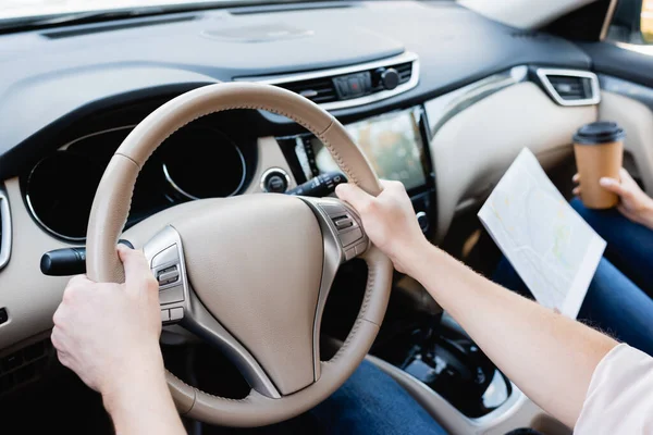 Vista recortada del hombre que conduce el coche cerca de la esposa con el mapa y el café para llevar en el fondo borroso - foto de stock