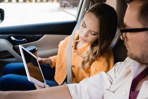 Woman with coffee to go showing digital tablet at husband driving car on blurred foreground — Stock Photo