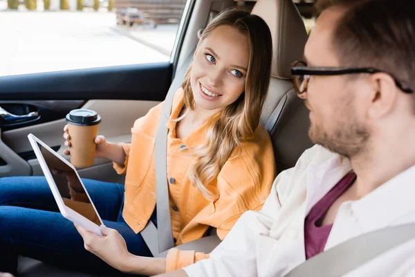 Smiling woman with coffee to go and digital tablet looking at husband on blurred foreground in car — Stock Photo