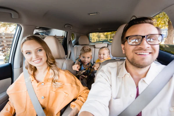 Cheerful kids pointing with fingers near parents on blurred foreground in car — Stock Photo