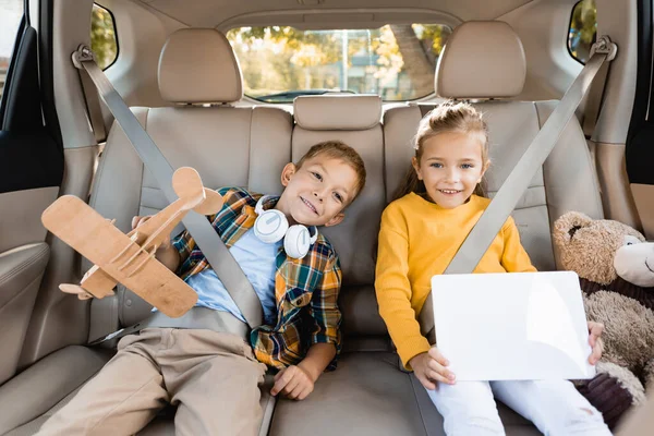 Веселі діти з іграшками та цифровим планшетом на задньому сидінні автомобіля — стокове фото