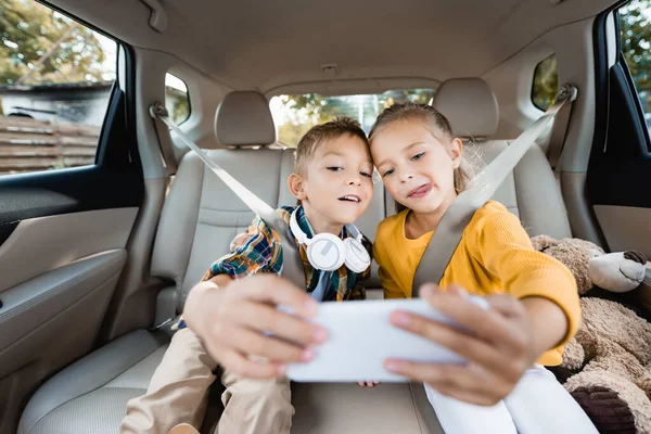 Children with headphones and toy taking selfie on smartphone on blurred foreground in car — Stock Photo