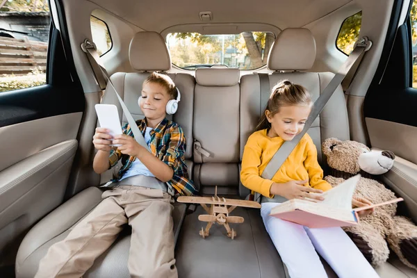 Усміхнені діти зі смартфоном і книгою сидять біля іграшок на задньому сидінні автомобіля — стокове фото