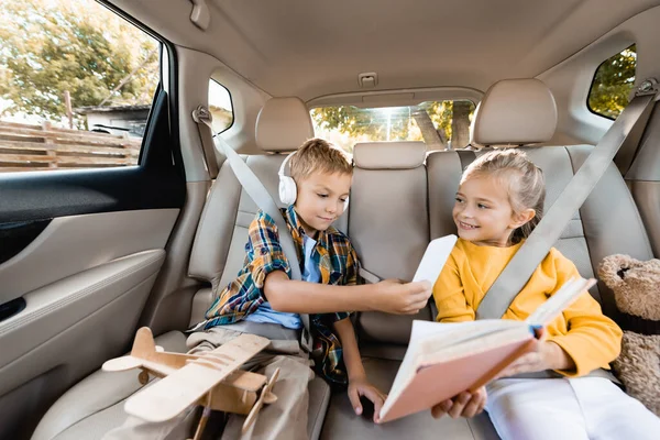 Niño en auriculares con teléfono inteligente cerca de la hermana sonriente con libro cerca de juguetes en el coche - foto de stock