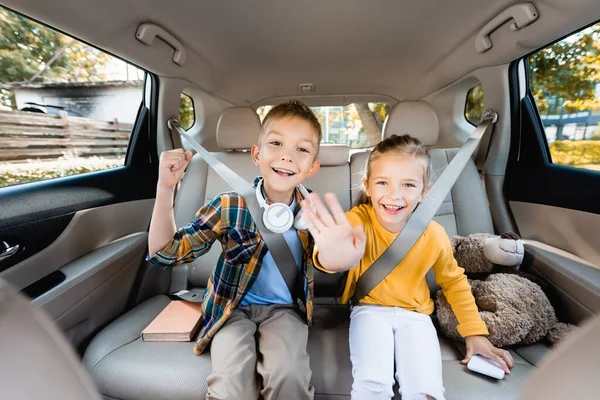 Souriant enfants montrant oui et agitant à la caméra près du smartphone et réserver en voiture — Photo de stock