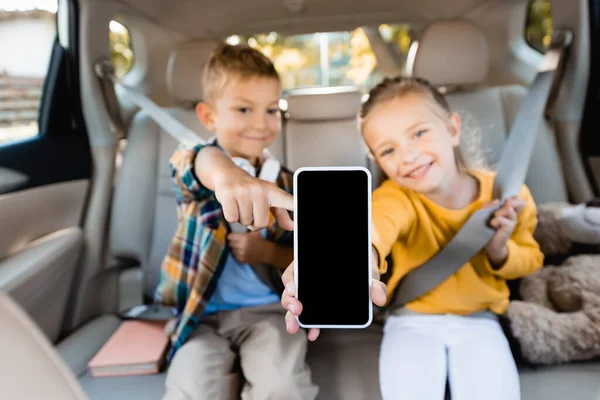 Smartphone con pantalla en blanco en la mano de los niños sonrientes sobre fondo borroso en el coche - foto de stock