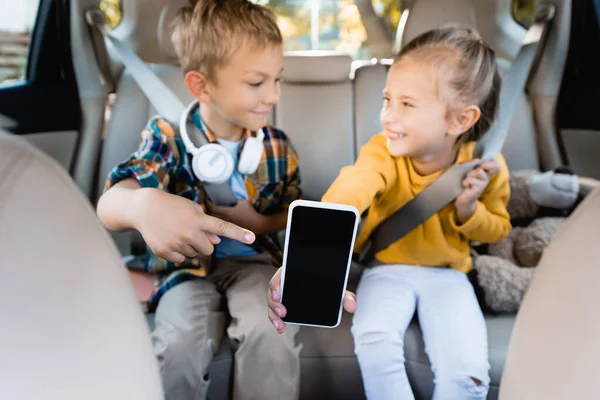 Smartphone con pantalla en blanco en la mano de niña sonriente y niño señalando con el dedo en auto sobre fondo borroso - foto de stock