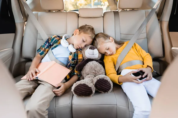 Niños con smartphone, libro y juguete suave durmiendo en auto durante el viaje - foto de stock