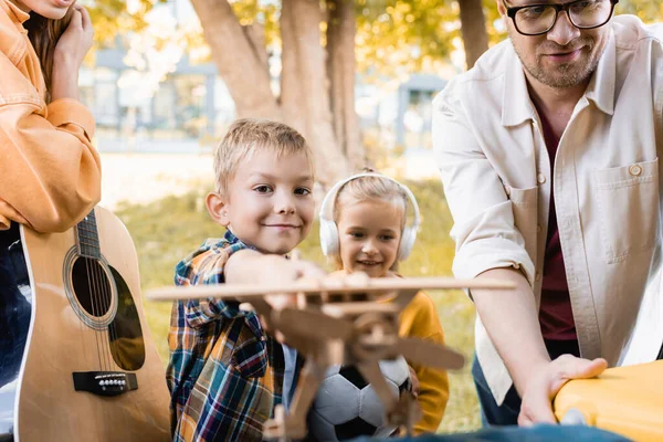 Boy holding toy plane near smiling sister in headphones and parents with acoustic guitar outdoors — Stock Photo