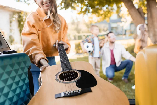 Vista cortada de mulher sorridente levando guitarra acústica do porta-malas do carro perto da família no fundo borrado ao ar livre — Fotografia de Stock