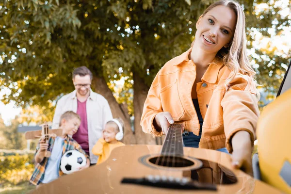 Lächelnde Frau mit Akustikgitarre aus dem Kofferraum neben Mann und Kindern auf verschwommenem Hintergrund im Freien — Stockfoto