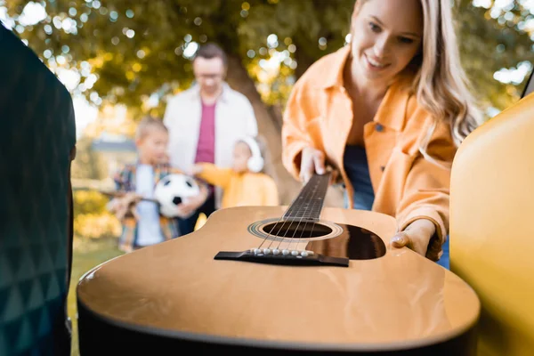 Guitare acoustique dans les mains d'une femme souriante près du coffre de la voiture et de sa famille sur fond flou — Photo de stock