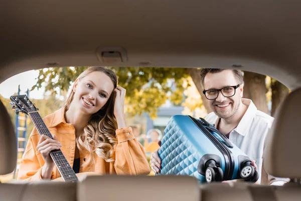 Lächelndes Paar mit Akustikgitarre und Koffer blickt in die Kamera in der Nähe des Kofferraums des Autos auf verschwommenem Vordergrund — Stockfoto