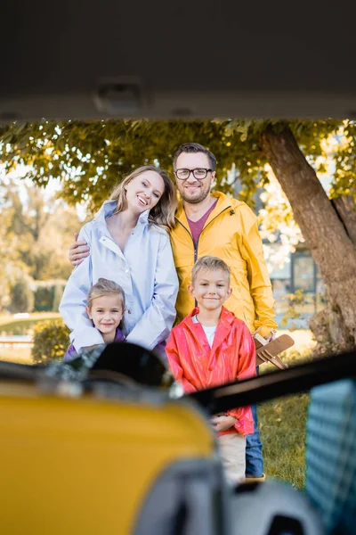 Lächelnde Familie blickt auf verschwommenem Vordergrund in die Kamera am Kofferraum des Autos — Stockfoto